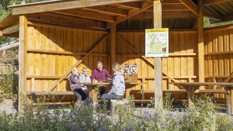 A cozy wooden accommodation with an outdoor seating area. Three people are sitting together and enjoying their time in the greenery.