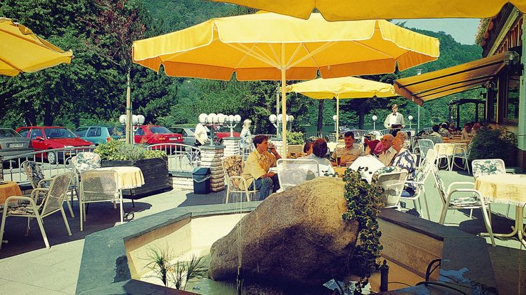 Een café in de openlucht met gele parasols en een kleine fontein. Gasten zitten aan tafels en genieten van de omgeving.