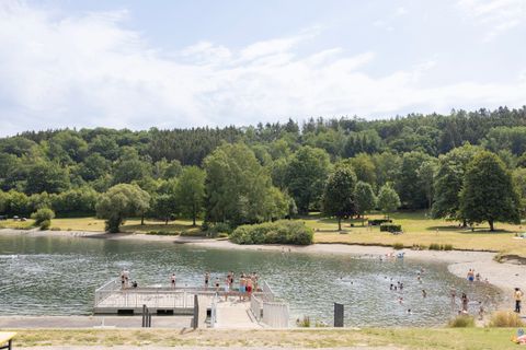 Ein ruhiger See umgeben von grünen Bäumen und Wiesen. Auf der Uferpromenade sind Menschen zu sehen, die sich entspannen und das Wasser genießen.