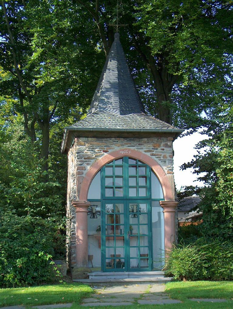Een historisch gebouw met een puntig dak en grote ramen. Omgeven door groene bomen en een verzorgde tuin.