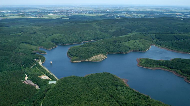 Een schilderachtig landschap met een rustige meer, omringd door dichte bossen. De lucht is helder en de natuur is weelderig groen.