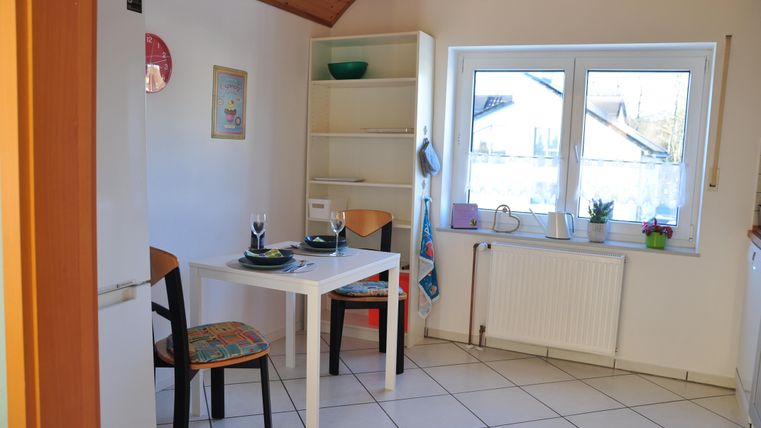 A bright kitchen with a small table and two chairs. In the background, there is a shelf and a window with a view outside.