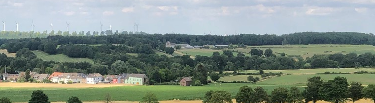 Eine weite, grüne Landschaft mit Hügeln und einem blauen Himmel. Im Vordergrund sind verstreute Häuser und Bäume zu sehen.