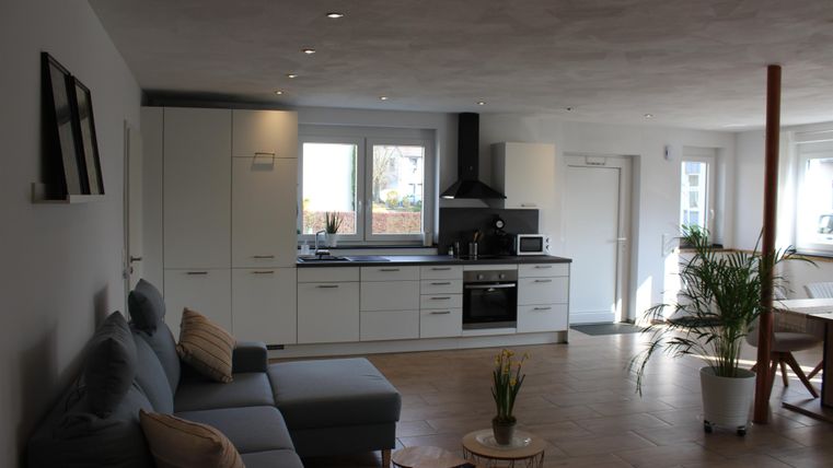 A modern, bright kitchen with an open living area. In the foreground is a cozy sofa and a small table with a plant.