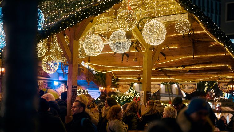 Ein festlich dekorierter Marktplatz mit Lichtern und schimmernden Kugeln. Viele Menschen genießen die Weihnachtsatmosphäre.