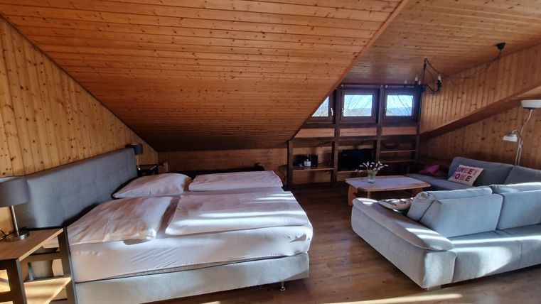 A cozy attic space with two beds and a sofa. The wooden paneling and large windows create an inviting atmosphere.