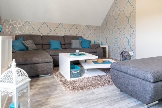 Ein modernes Wohnzimmer mit einer grauen Couch und blauen Kissen. Der Tisch ist weiß und es gibt dekorative Elemente wie einen Teppich und eine Laterne.