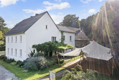 Ein weißes Haus umgeben von grüner Vegetation. Im Vordergrund befindet sich ein Zelt, das für Veranstaltungen genutzt werden kann.