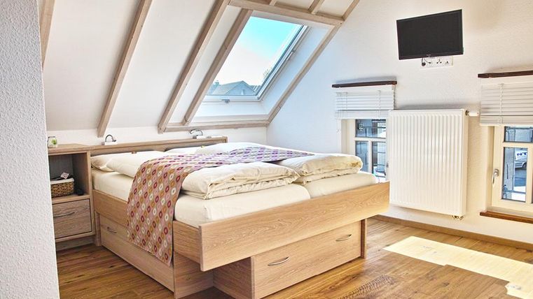 A cozy bedroom with a double bed and wooden floor. The room has sloping walls, a window with a view, and a television on the wall.
