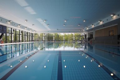 Ein modernes Hallenbad mit einem klaren Wasserbecken. Große Fenster lassen viel Tageslicht herein und bieten einen Blick auf die Außenanlage.