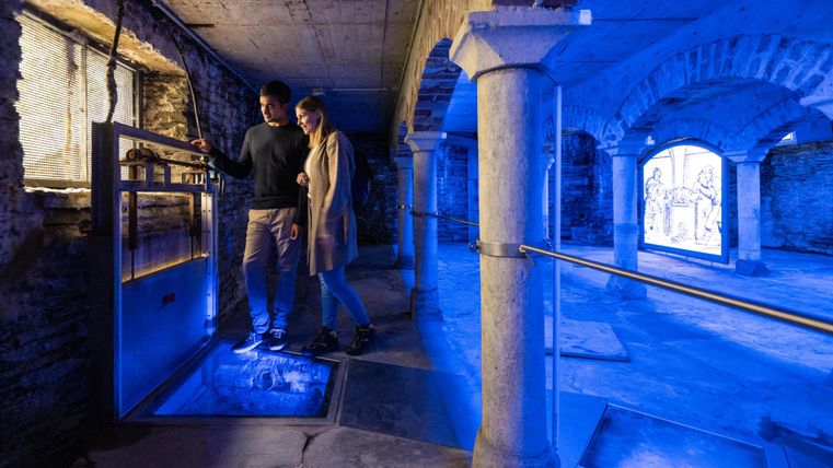 A modern basement with blue lighting. Two people are exploring the space, which is designed with columns and windows.