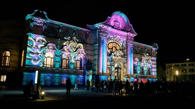Ein historisches Gebäude, das nachts bunt beleuchtet ist. Viele Menschen stehen davor und bewundern die Lichtshow.