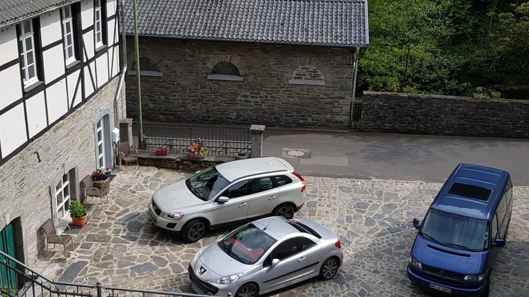 Een geplaveide binnenplaats met drie auto's en een historisch gebouw op de achtergrond. De omgeving is groen en rustig.