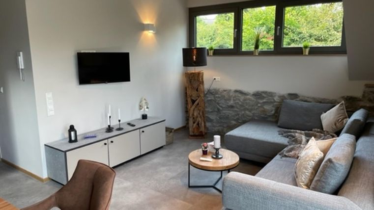 Een moderne woonkamer met een grijze bank en een tafel. Grote ramen zorgen voor veel daglicht en uitzicht op groen.