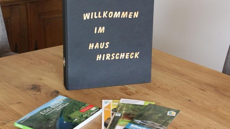 A black folder with the inscription "Welcome to the Hirschcheck House" is lying on a wooden table. Next to it are several brochures about the surroundings.