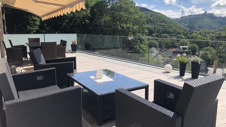 A modern terrace featuring a black rattan seating area and a glass table. In the background, green hills and a bright blue sky are visible.