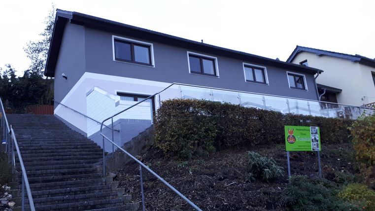 A modern building with a grey facade and large windows, surrounded by a landscaped area. The staircase leads to the main entrance of the house.