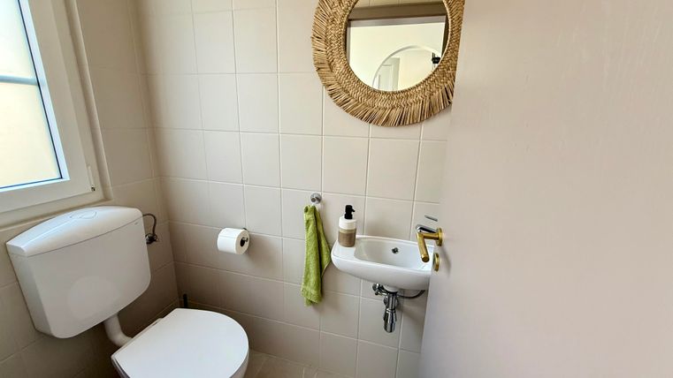 Een kleine, lichte badkamer met een toilet en een wastafel. Een ronde spiegel van natuurlijk materiaal hangt aan de muur.