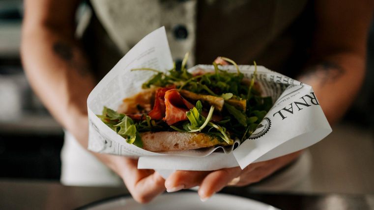 A freshly prepared sandwich with ham and salad, wrapped in paper. It is held by a person standing at a table.