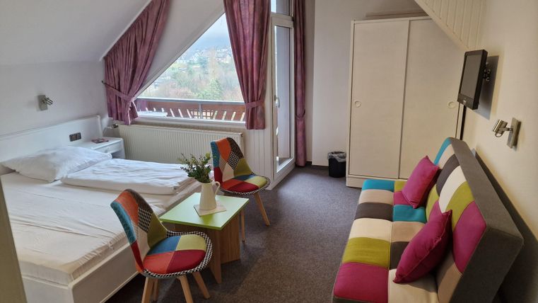A bright room with a window and a beautiful view. There is a bed, a table with colorful chairs, and a sofa.