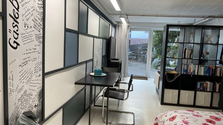 Een moderne kamer met een tafel en stoelen en planken voor boeken. Grote ramen zorgen voor veel daglicht.