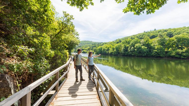 A hiking trail by a quiet river surrounded by lush greenery. Two people are exploring nature and enjoying the view.