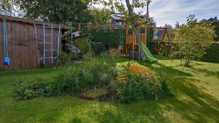 Ein schöner Garten mit bunten Blumen und einer Rutsche für Kinder. Im Hintergrund sind ein Holzschuppen und ein Spielgerüst zu sehen.