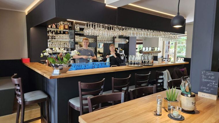 A modern bar with a wooden counter and numerous glasses. Two employees stand friendly behind the bar.