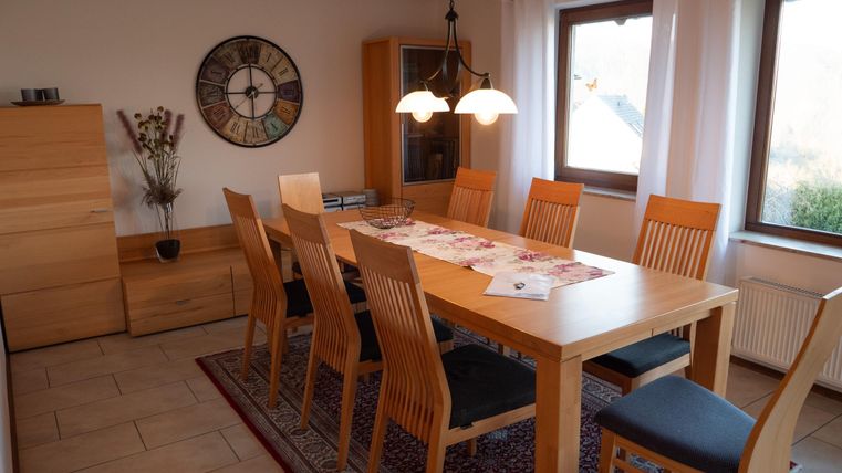 A bright dining area with a large wooden table and ten chairs. Windows and a round wall calendar hang on the walls.