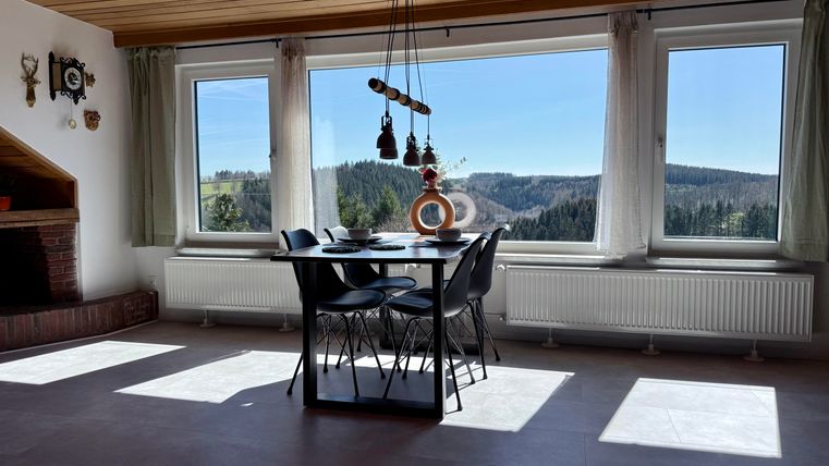 Een moderne eetkamer met een tafel en stoelen, omgeven door grote ramen. Door de ramen heeft men een prachtig uitzicht op het bosrijke landschap.