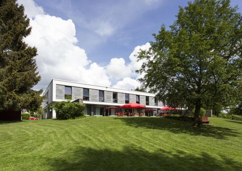 A modern building surrounded by green grass and trees. Red sun umbrellas are on the terrace.