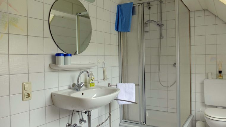 A bright bathroom with white tiles. It features a sink, a shower, and a toilet.