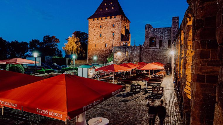 A cozy outdoor restaurant with red umbrellas and a historic castle in the background. The scene is lit at night and conveys an inviting atmosphere.