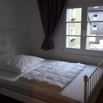 A simple bedroom with a white bed and a window. The walls are light, and there are curtains that gently diffuse the light.