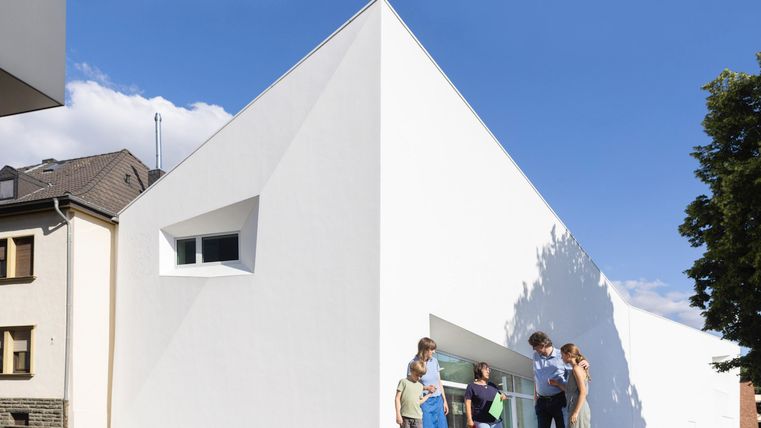 Ein modernes, weißes Gebäude mit einer markanten, dreieckigen Form. Vor dem Gebäude stehen mehrere Personen, die miteinander interagieren.