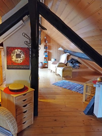 Een gezellige zolderkamer met houten plafonds en een warme ambiance. De inrichting bestaat uit een rotanstoel, planken en een rustieke commode.