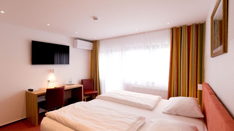 A modern hotel room with a double bed and a television on the wall. The room has large windows with colored curtains and a small workspace.