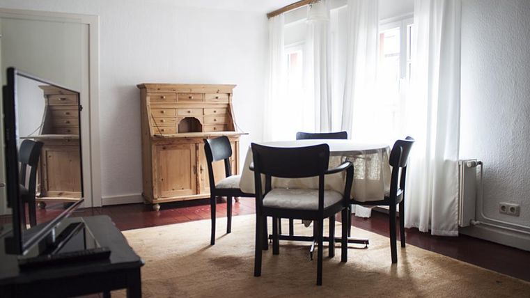 Een lichte kamer met een tafel en stoelen. Op de achtergrond staat een elegante kast en grote ramen laten veel licht binnen.