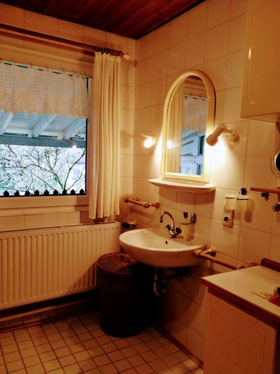 Een kleine badkamer met een wastafel, een spiegel en een raam. De muren zijn licht betegeld en er is een warme sfeer door de verlichting.