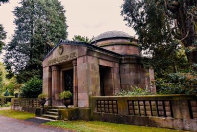 Ein historisches Mausoleum aus Stein, umgeben von Bäumen und grünem Gras. Die Architektur zeigt klare Linien und einen runden Dachaufsatz.