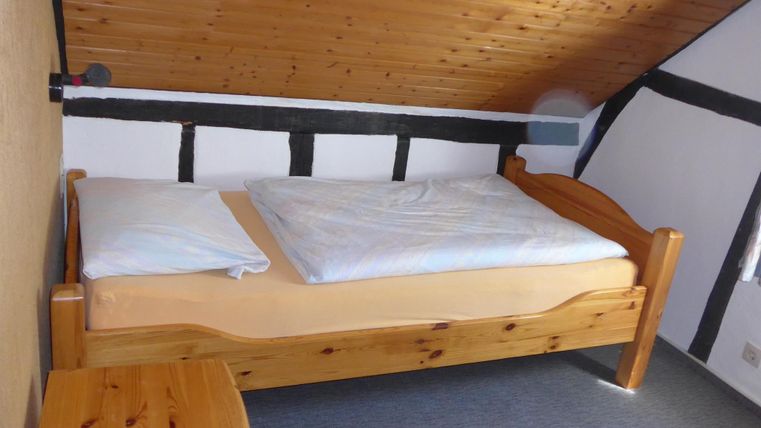 A simple bedroom with a wooden bed and beige bedding. The walls are partially paneled with wood and the ceiling is slanted.