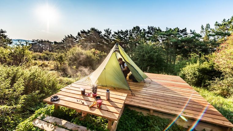 Een tent op een houten platform te midden van groene natuur. De zon schijnt en de omgeving is rustig en uitnodigend.