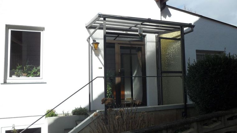 Een modern entreeportaal met een glazen overkapping. De gevel van het huis is wit en heeft grote ramen.