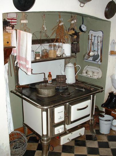 A nostalgic kitchen with an old wooden stove, shelves, and various kitchen utensils. Dried herbs and additional cooking tools are hanging on the wall.