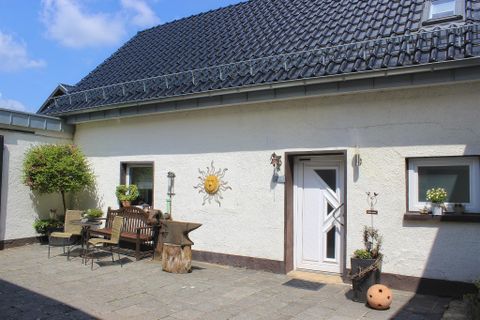 Een rustige terras met houten bank en tafel, omringd door planten. Het huis heeft een lichte gevel en een zonnig wandbord.
