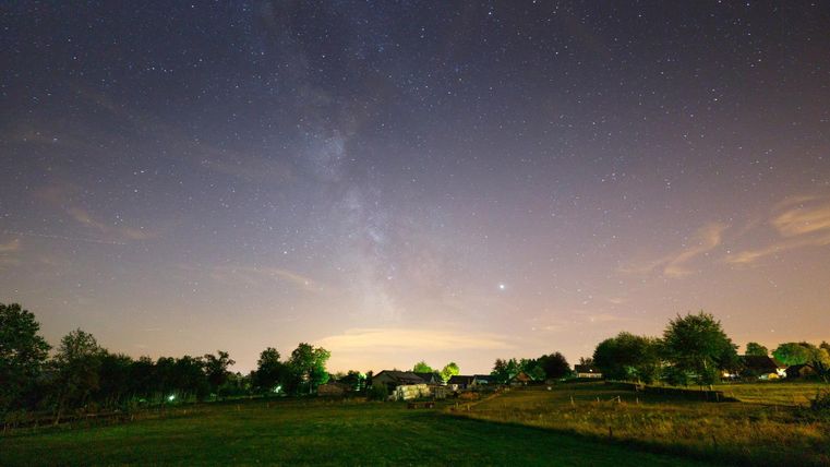 A clear night sky full of stars and the Milky Way are visible. In the foreground lies a tranquil landscape with trees and gentle hills.