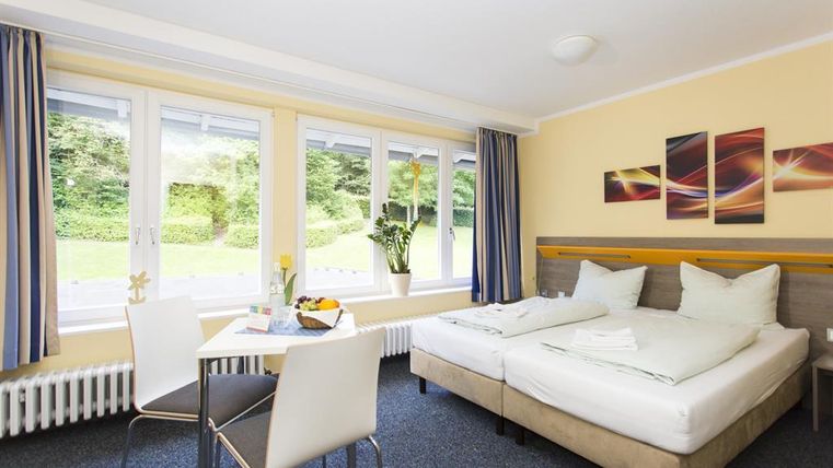 A bright hotel room with two beds, a table and chairs. Large windows provide plenty of daylight and a view of greenery.