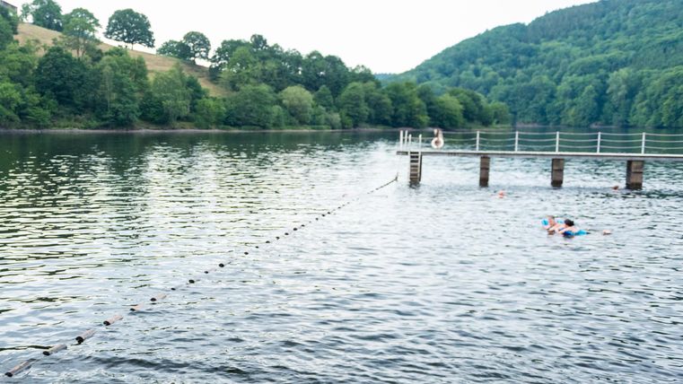 Een rustige vijver met groene heuvels op de achtergrond. Op het water drijft een persoon dicht bij een houten steiger.