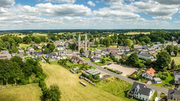 Eine malerische Landschaft mit einem kleinen Dorf und einer Kirche im Hintergrund. Umgeben von grünen Feldern und Bäumen bietet die Szene eine ruhige Atmosphäre.