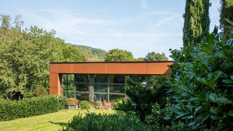 Een modern huis met grote ramen te midden van een groene landschap. Omgeven door bomen en een verzorgde tuin.